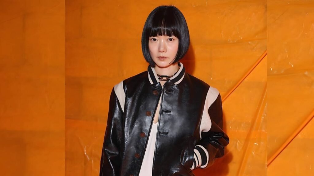 Bae Doona ( 배두나 ) with a short black bob stands before an orange backdrop, wearing a black glossy varsity jacket with white striped cuffs