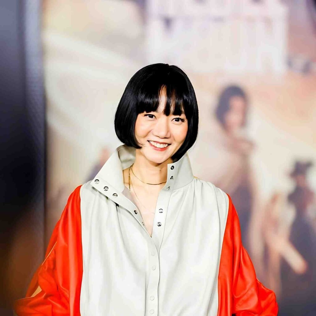 Bae Doona with a sleek black bob and bangs, wearing a pale blouse with a studded collar and red sleeves.