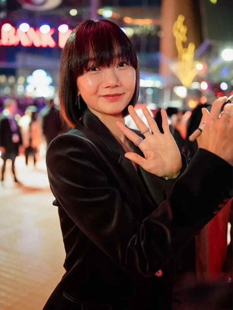 Bae Doona boyfriend with a short bob in a black blazer waves at the camera; neon night, blurred crowd, and rings on her fingers.