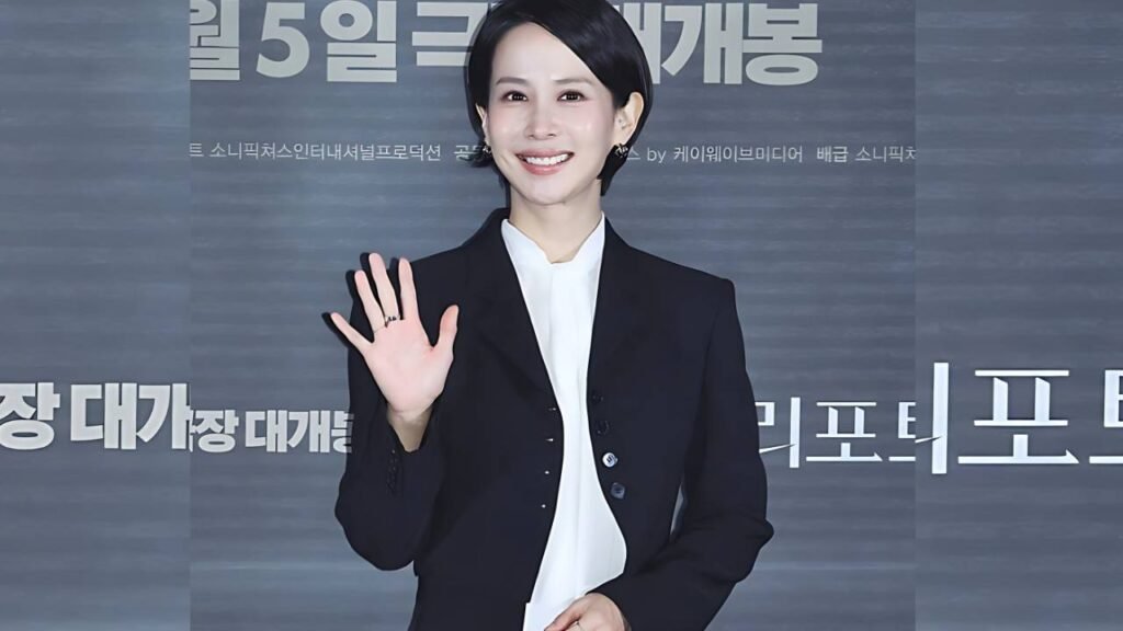Cho Yeo-jeong in a black blazer and white blouse waves to the camera at a press event, standing before a gray backdrop with large Korean text.