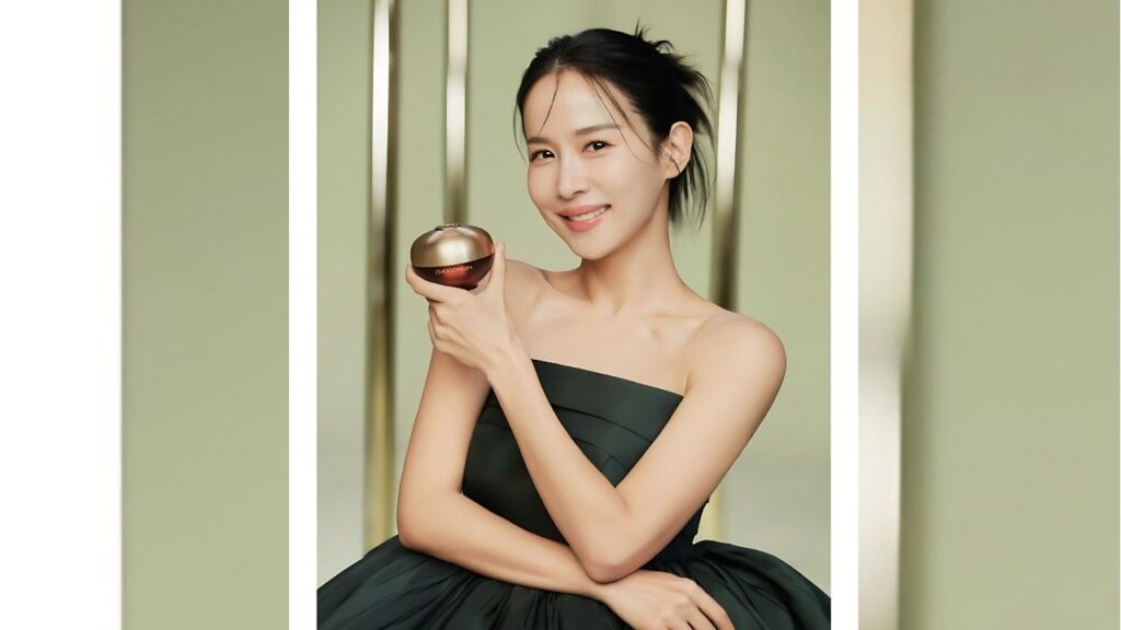Cho Yeo-jeong in a strapless dark green gown stands between green panels, holding a round brown cosmetic compact and smiling.