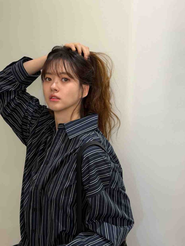 Go Ara with a messy ponytail lifts her hair with one hand, wearing a navy striped oversized shirt against a pale wall