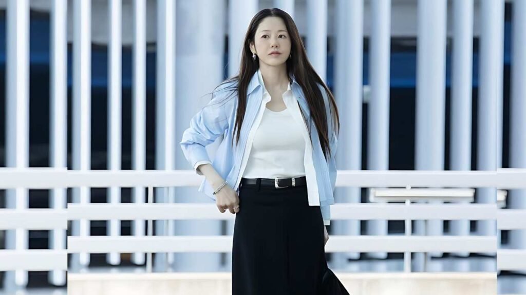 Go Hyun Jung (고현정) with long dark hair stands on a pale blue and white bridge, wearing a light blue shirt over a white top and black pants, hand on hip.
