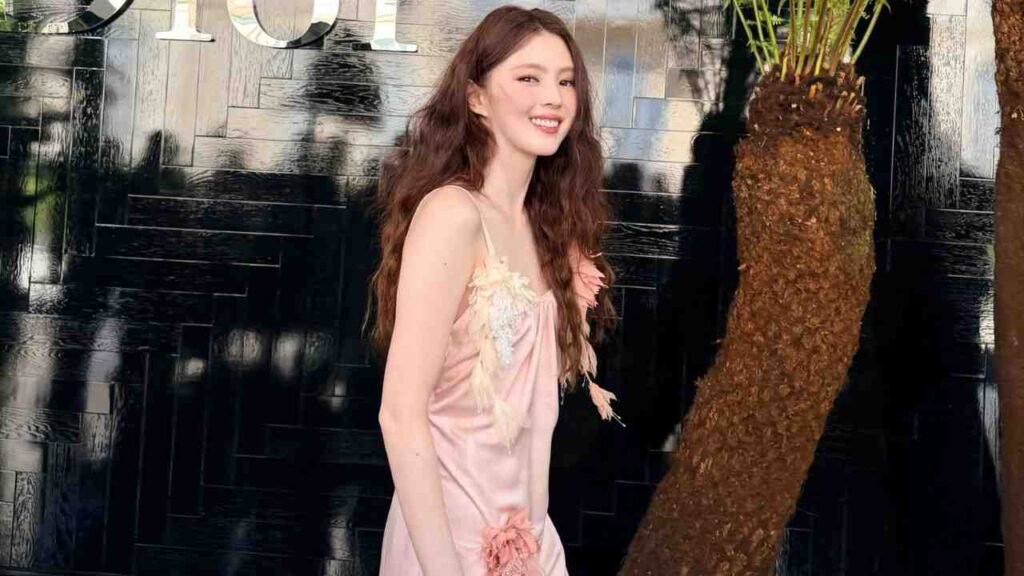 Han So-hee ( 한소희 ) with wavy brown hair in a satin slip dress, standing beside a palm trunk against a glossy black wall.