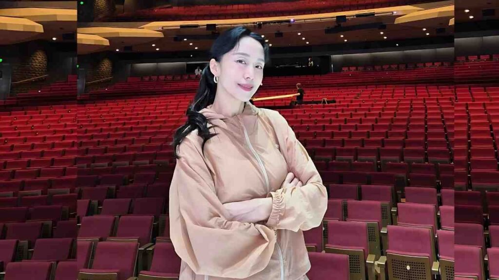 Jeon Do-yeon ( 전도연 ) with black hair and hoop earrings stands with crossed arms in an empty theater, wearing a peach jacket, seats behind.