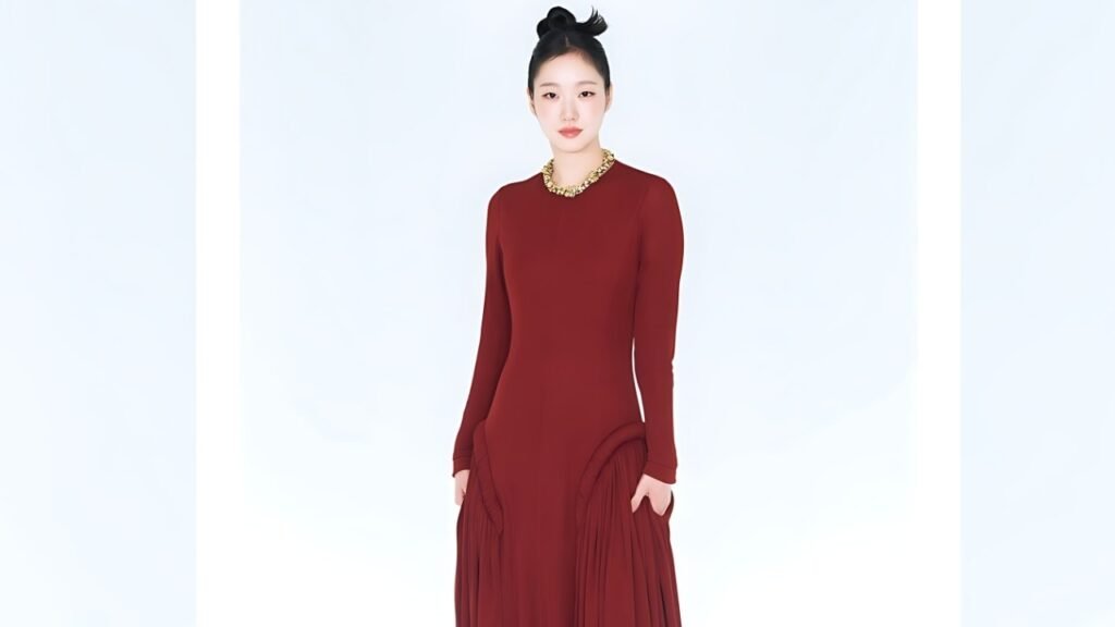 Kim Go-eun in a burgundy long-sleeve gown with side draped pleats, gold necklace, hair in a top knot, against backdrop.