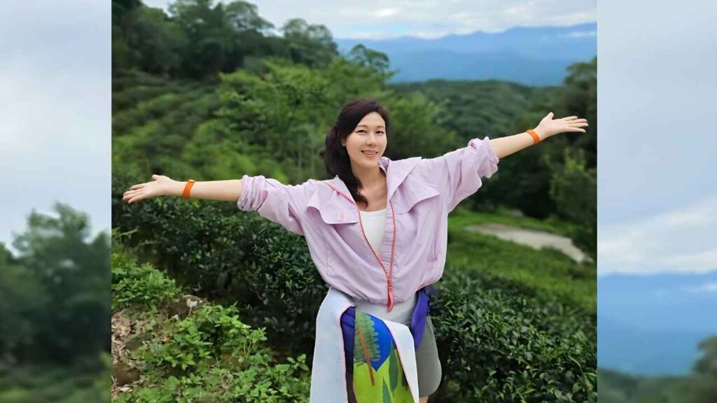 Kim Ha-neul on a green hillside with arms outstretched, wearing a light pink jacket over a white top and orange bracelets.