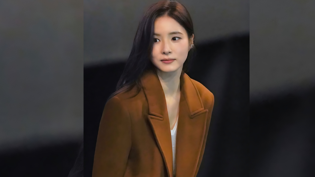 Kim Hye-soo ( 김혜수 ) with long dark hair wearing a rust-brown coat over a white top, gazing softly to the left against a dark blurred background.