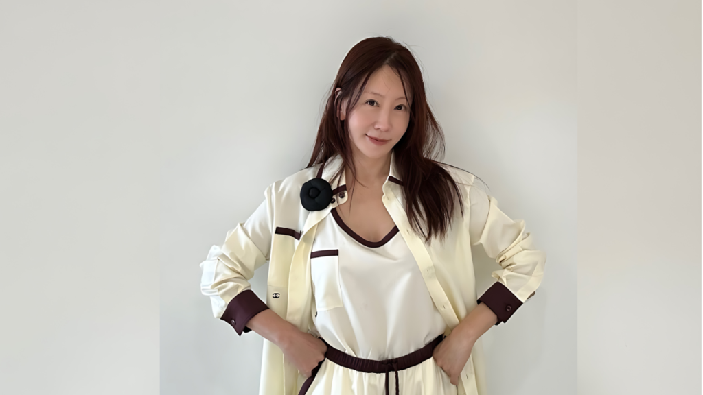 Kim Jung-eun ( 김정은 ) with long dark hair in a cream outfit poses with hands on hips against a pale wall, a black flower pin at collar.