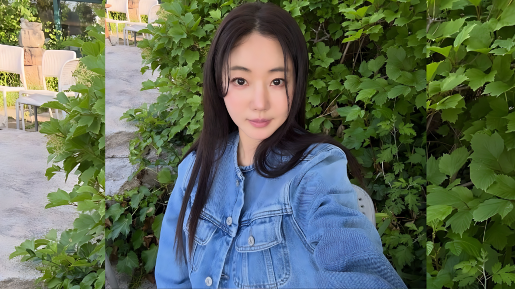 Kim Sa-rang ( 김사랑 ) with long dark hair in a blue denim jacket posing for a selfie, lush green vines and white chairs behind.