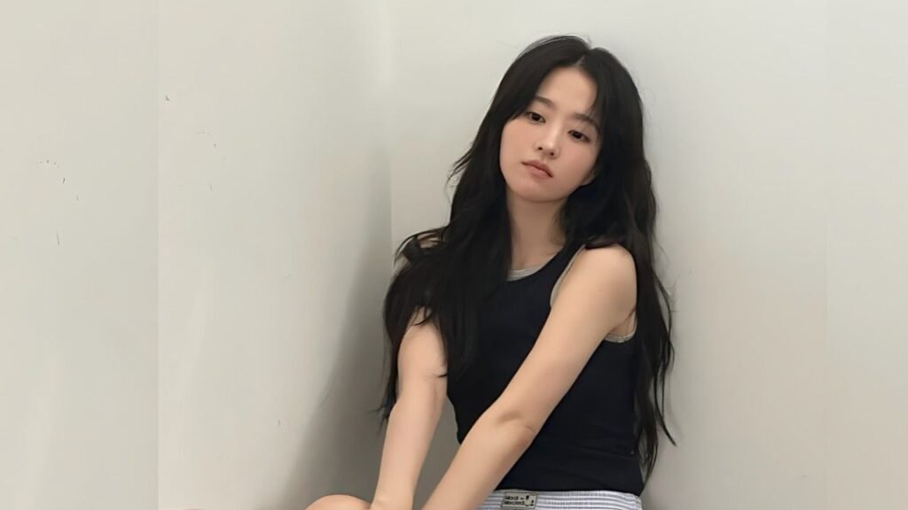 Kim So-hyun (김소현) with long black hair sits against a pale wall, wearing a black sleeveless top and light striped pants, calm.