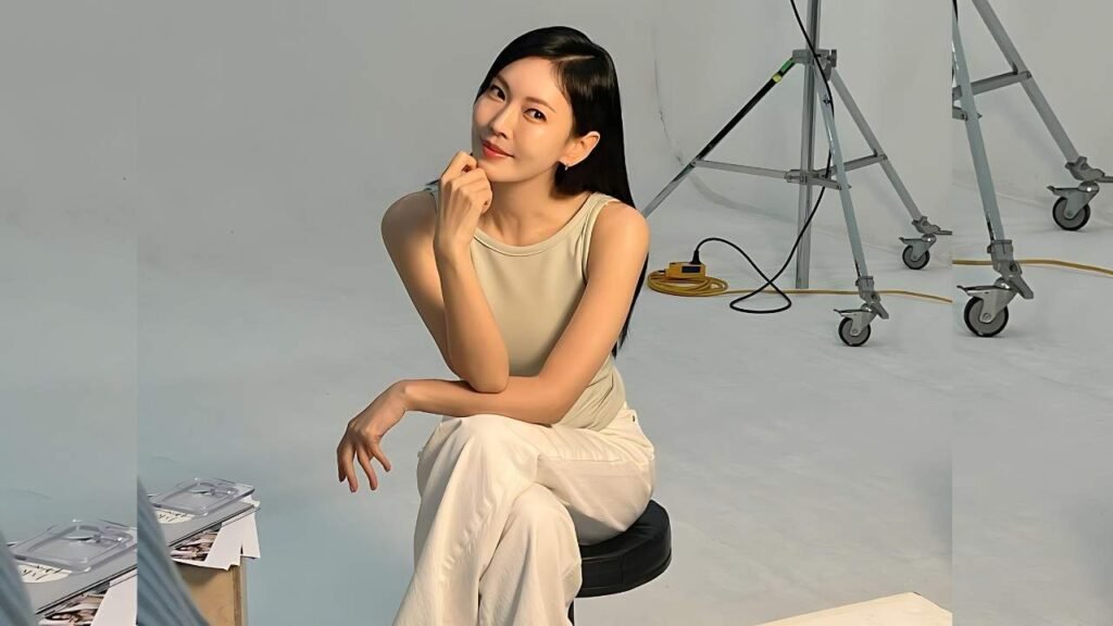 Kim So-yeon ( 김소연 ) sits on a black stool in a beige sleeveless top and light pants, chin on her hand in a studio with ladders and cables