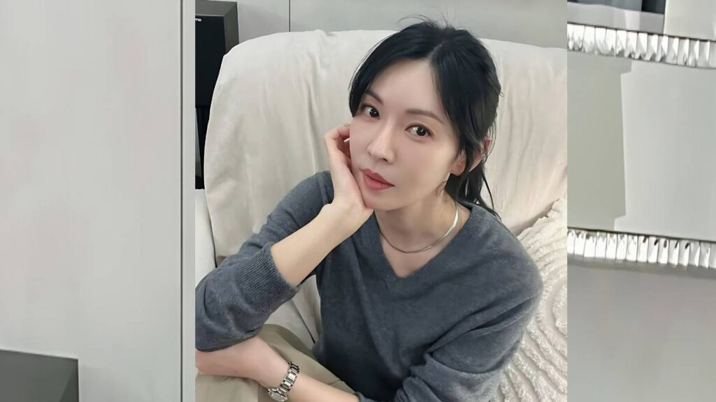 Kim So-yeon with dark hair in a gray sweater sits on a beige couch, chin resting on her hand, wearing a silver necklace.