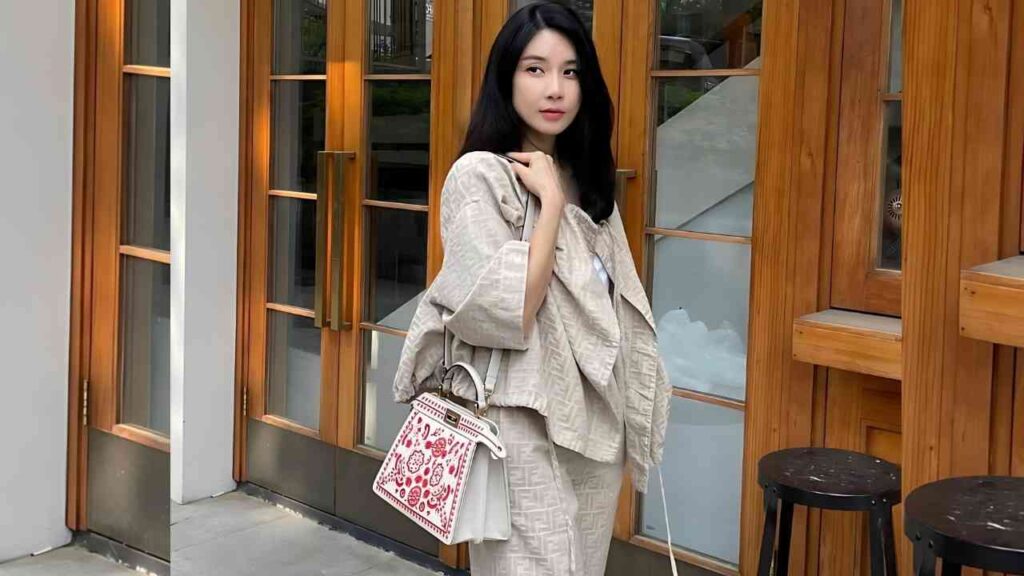 Lee Bo-young with black hair in a beige outfit stands outdoors by wooden doors, carrying a white handbag with red embroidery.