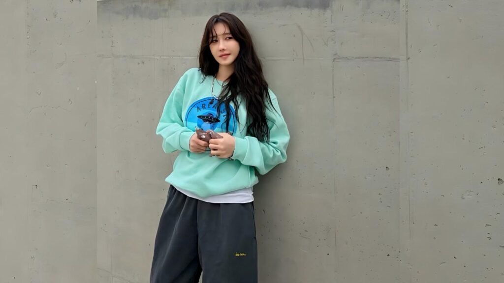 Lee Ji-ah (이지아 ) with long dark hair leans against a gray concrete wall, wearing a mint green oversized sweatshirt, holding phone.