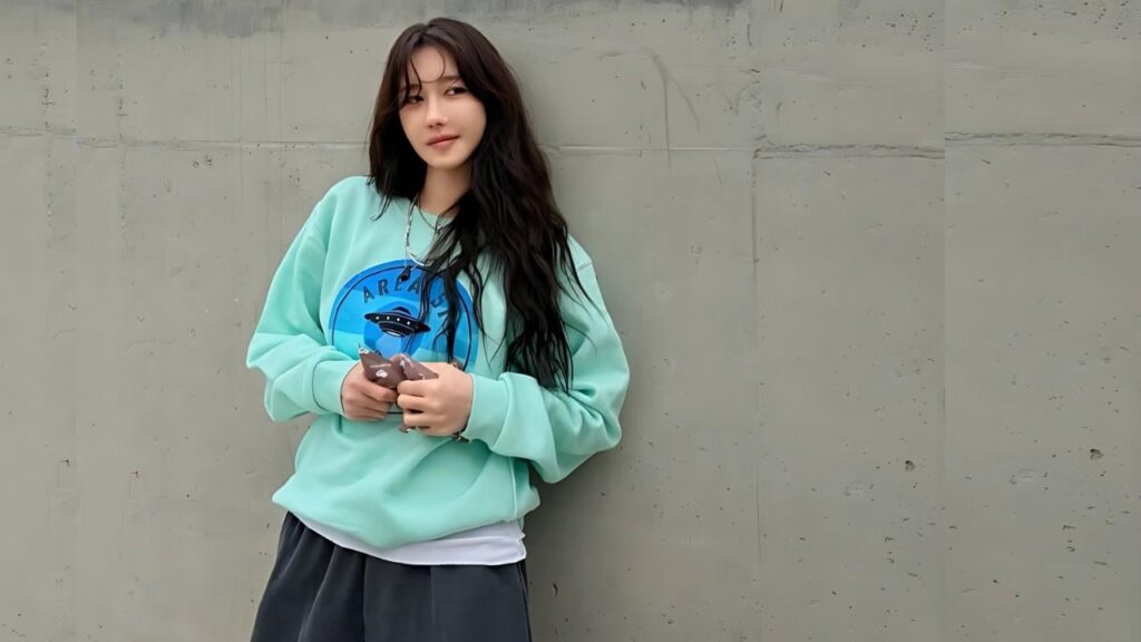 Lee Ji-ah with long dark hair leans against a gray concrete wall, wearing a mint sweatshirt with a blue circular graphic.