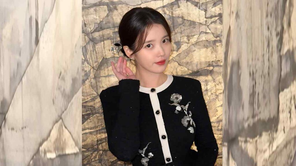 Lee Ji-eun in a black cardigan with white trim and floral embroidery poses against a marble wall, hand near ear, soft smile.
