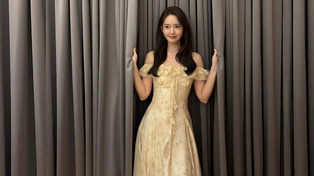 Lim Yoona ( 임윤아 ) in an ivory off-shoulder ruffled gown stands between dark pleated curtains, smiling softly.