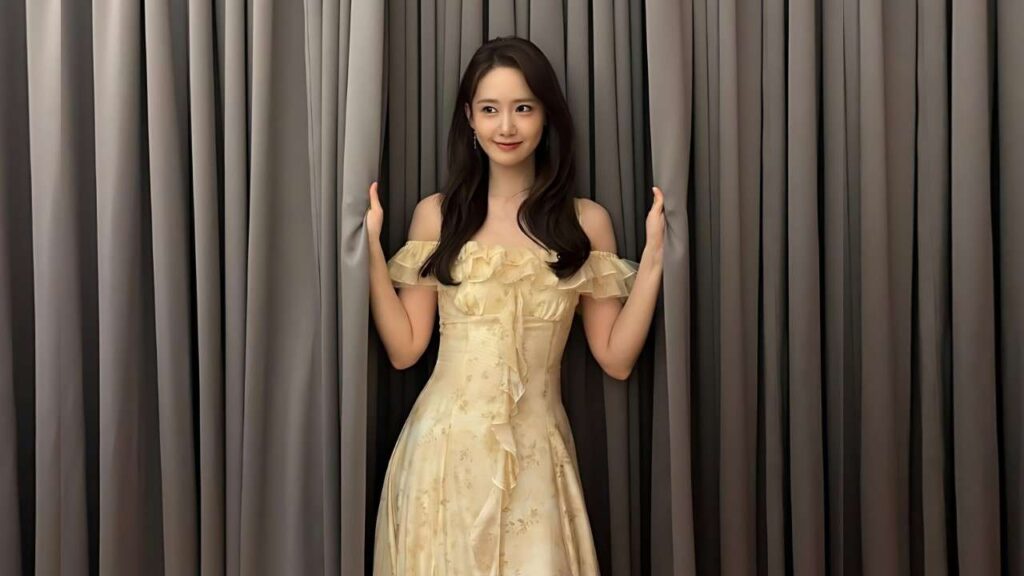 Lim Yoona in pale yellow off-shoulder dress with ruffles stands between dark gray curtains, hands on the panels, smiling softly.