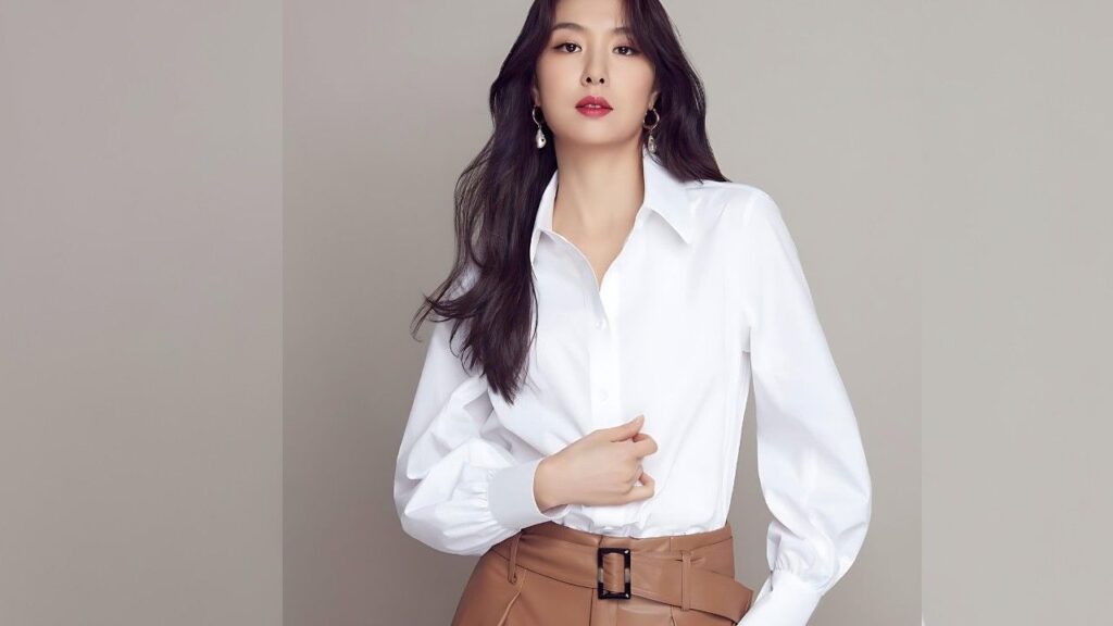 Seo Ji-hye in a white button-down blouse and tan belted pants poses against a beige studio backdrop, dark hair flowing, wearing drop earrings.