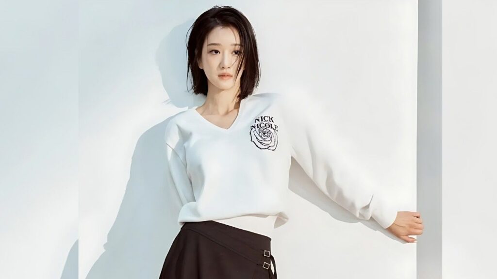 Seo Yea-ji stands against a white wall in a white V-neck sweatshirt with a rose emblem and a black belted skirt, gazing.