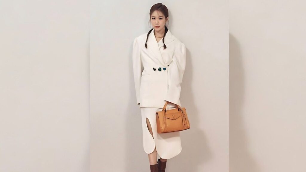 Yoo In-na in white oversized-shoulder blazer dress with curved hem and green buttons, carrying a tan leather handbag against beige backdrop.