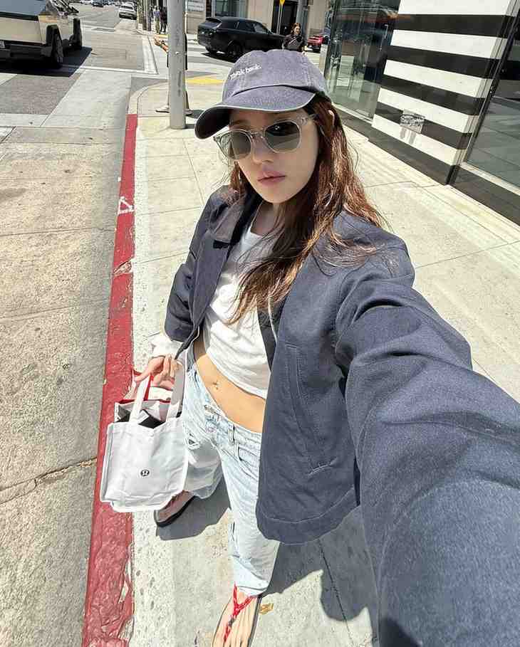 lee ji-ah height on a sunny city sidewalk takes a selfie, wearing a cap, sunglasses, navy jacket, white cropped top, light jeans, and a white tote