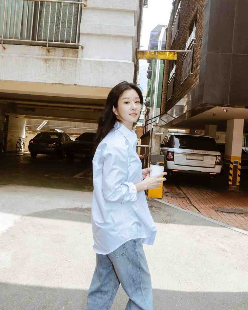 seo ye-ji in a pale blue oversized shirt and jeans walks through a sunlit urban alley, holding a coffee cup.