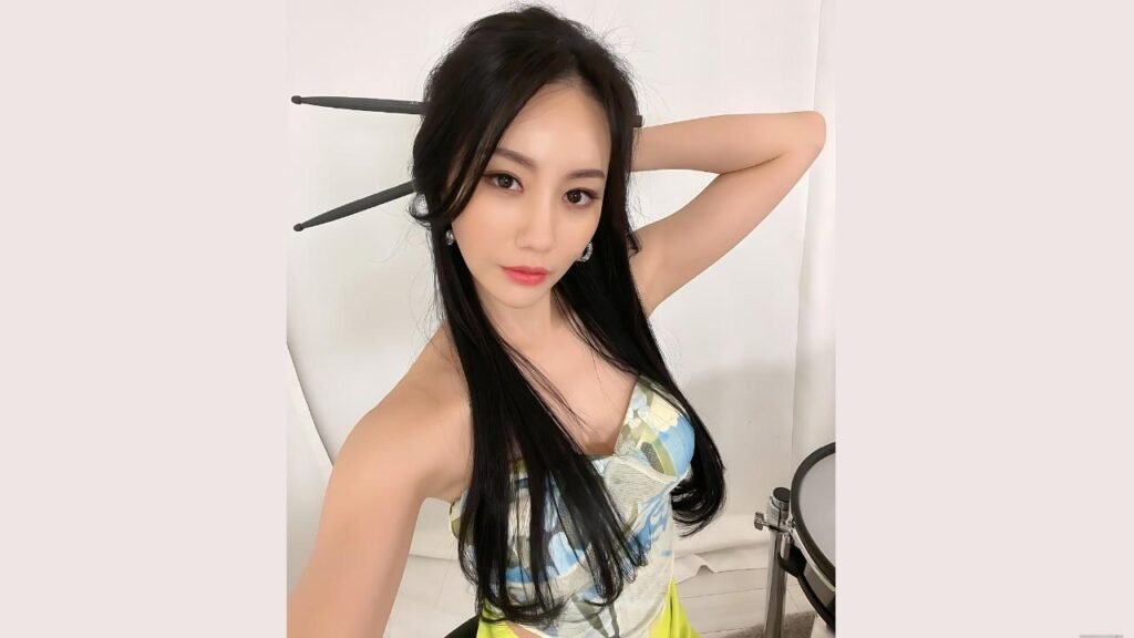 A-YEON with long black hair wears a patterned blue top, arm raised behind head, posing against a white backdrop.