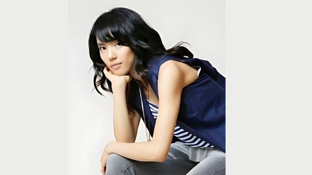 Ahn Gyu-ryun (안규련) with dark hair leans forward, chin on her hand, wearing a navy sleeveless top over striped tee and gray jeans.