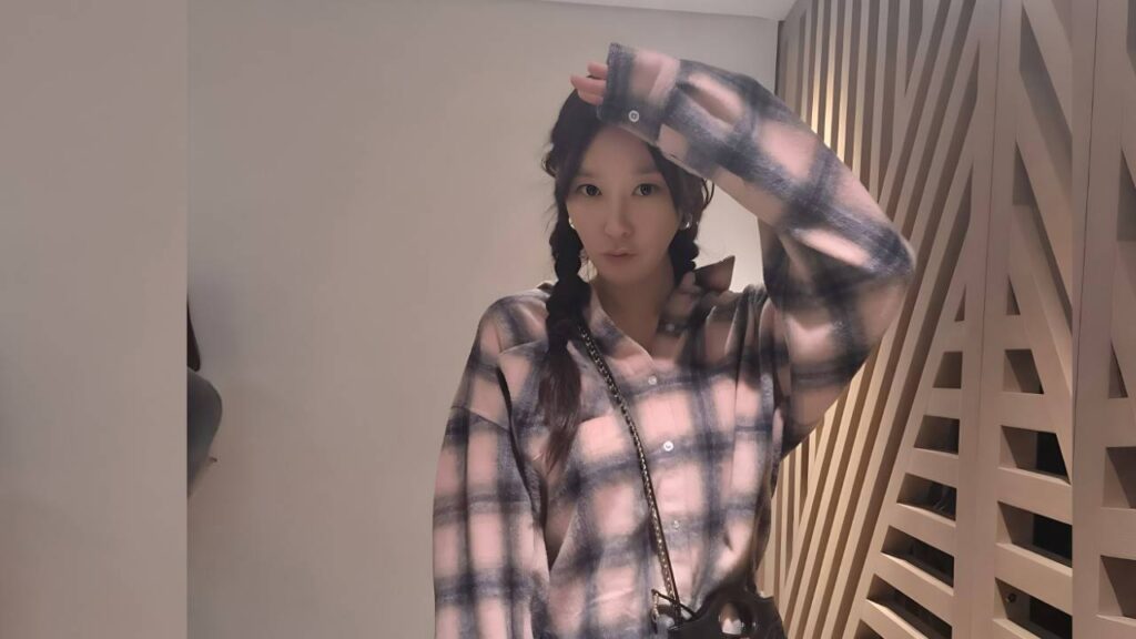 Bae Jin-ah (배진아) with braided pigtails in a pink-gray plaid shirt, raising her arm, beside a slatted wooden partition.