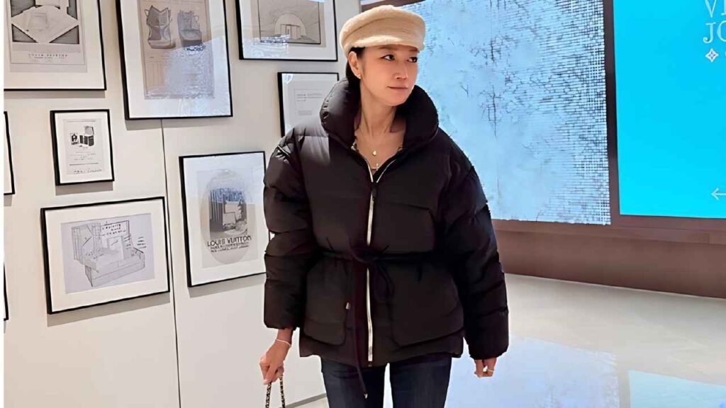 Byun Jung-soo in an oversized black puffer jacket and beige cap walks past framed sketches on a gallery wall, holding a chain-strap bag as a blue digital screen glows to the right.