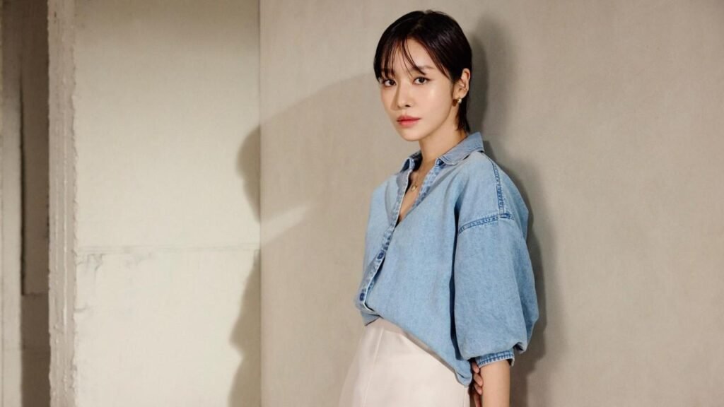 Cha Joo-young with short dark hair leans against a beige wall, wearing a loose denim shirt and pale skirt, calm expression.