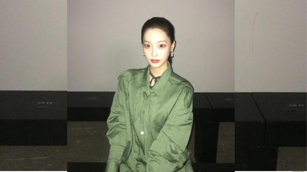 Choi Hee-jin seated against a pale wall, wearing an olive-green oversized jacket and neck scarf, with slicked-back hair.