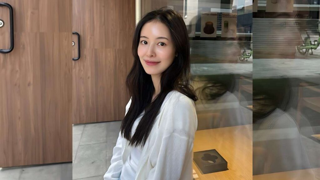 Choi Ji-woo (Ji Woo / 지우) with long dark hair sits indoors beside wood-paneled walls, in a white top, smiling softly.