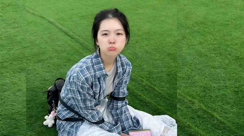 Choi Myung-bin sits on bright green turf wearing a blue plaid shirt over a gray tee, with a backpack and plush charms beside her.