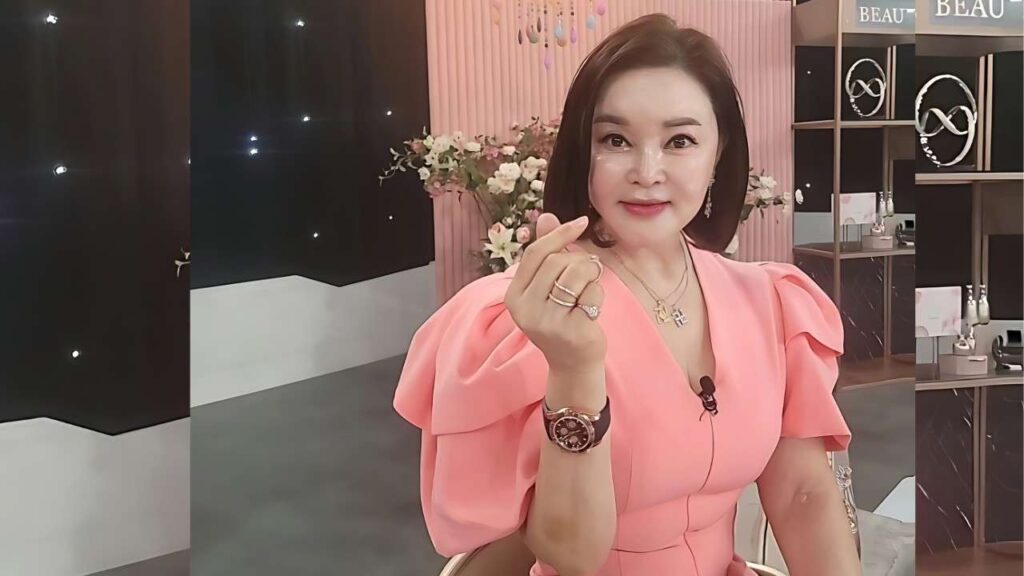 Choi Ran in a pink puff-sleeve dress sits in a studio, making a finger-heart gesture, wearing a watch, rings, and a necklace, with pink decor and jewelry displays behind