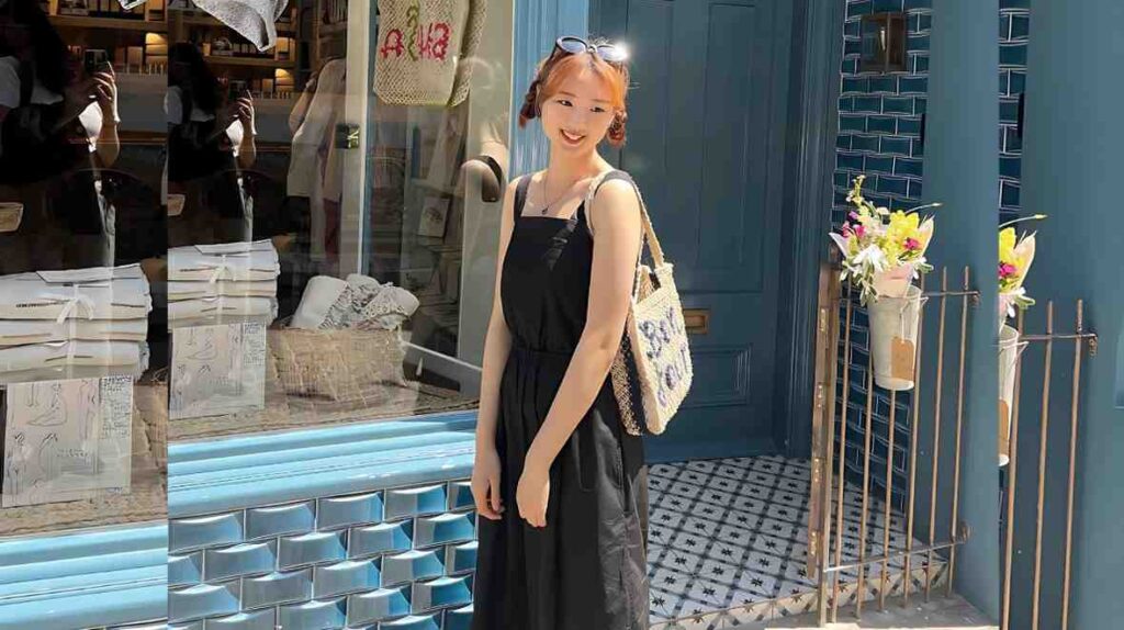 Choi Yu-hwa (최유화) in a black sundress stands outside a blue-tiled storefront, with a woven tote and glass reflecting the scene.