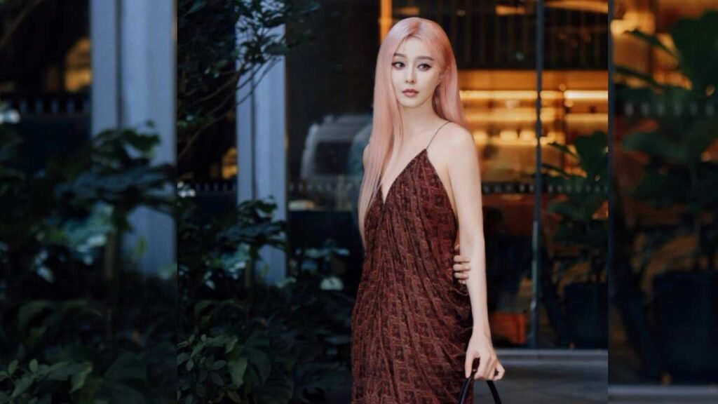 Fan Bingbing with long pastel pink hair in a brown patterned slip dress stands amid greenery, holding a bag and gazing ahead.