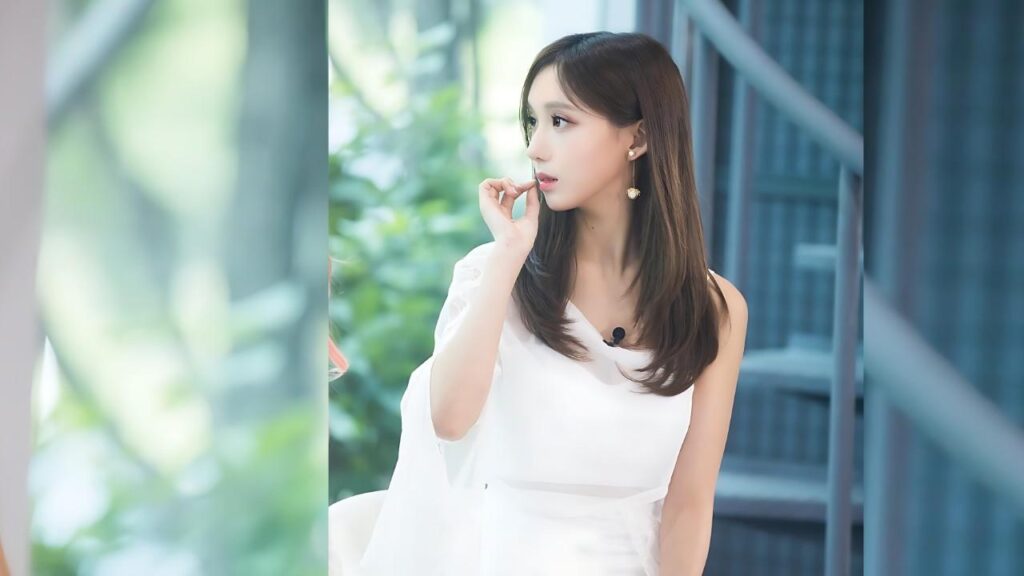 Go Doo-shim in a white dress stands by glass wall, hand to her lips, mic clipped, with greenery outside and blue.