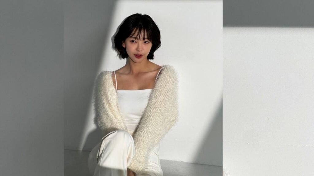 Go Won-hee with short black hair sits against a white wall, wearing a cream fluffy cardigan over a white top, in soft light.