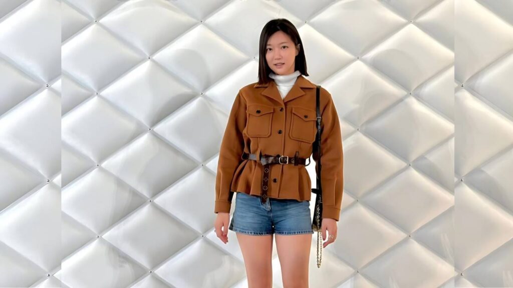 Ha Yeon-joo poses in front of a wall, wearing a tan belted jacket over a white turtleneck, denim shorts and a black bag.