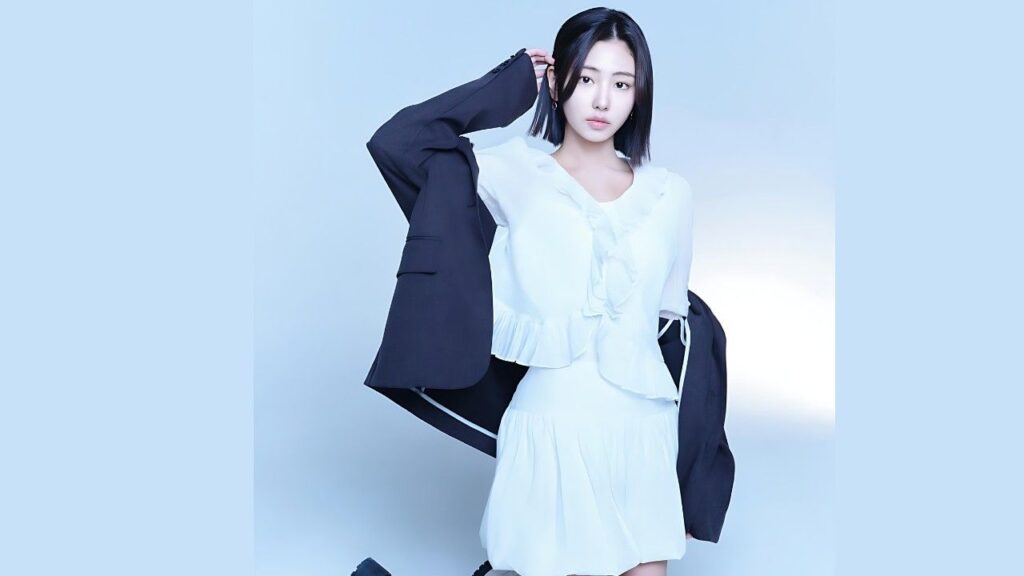 Ha Yul-ri (하율리) in a white ruffled dress with a dark blazer draped over her shoulders, against a pale blue studio backdrop.