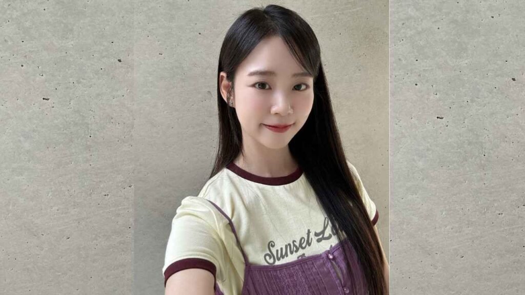 Han Seung-yeon with long black hair smiles for a selfie, wearing a cream shirt with maroon trim and purple corduroy pinafore.