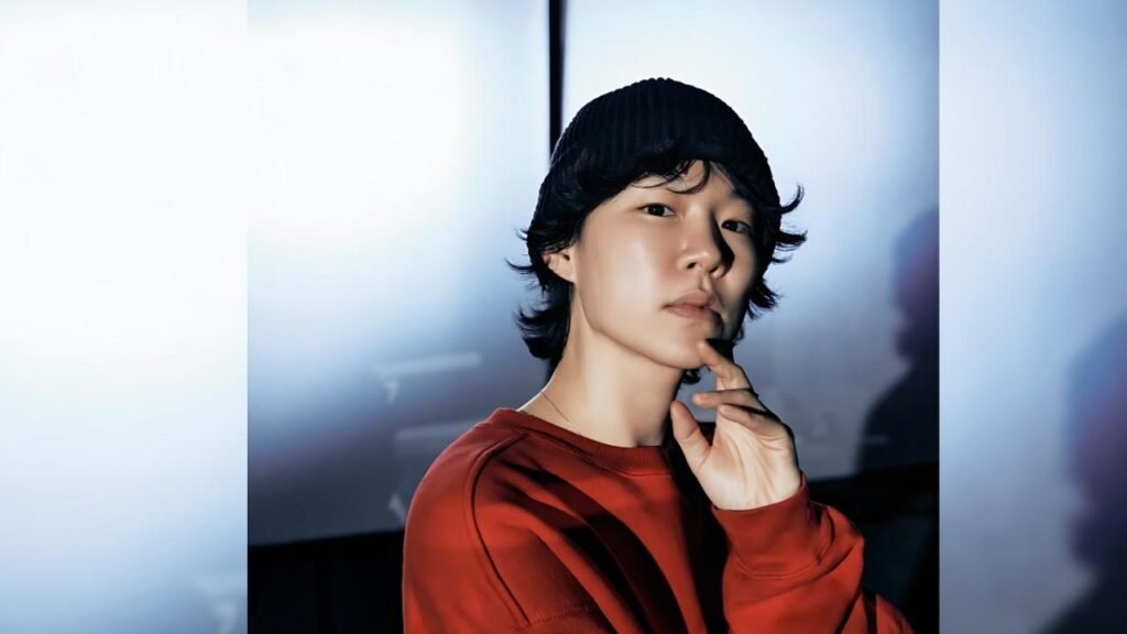 Han Ye-ri wearing a black beanie and red sweatshirt poses pensively, finger at lips, against a blue-lit studio backdrop.