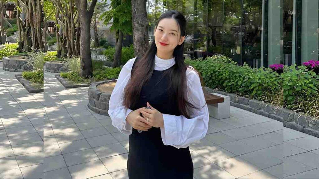 Hwang Sun-hee with long dark hair in a white puff-sleeve blouse over a black dress, hands clasped in a sunlit courtyard.
