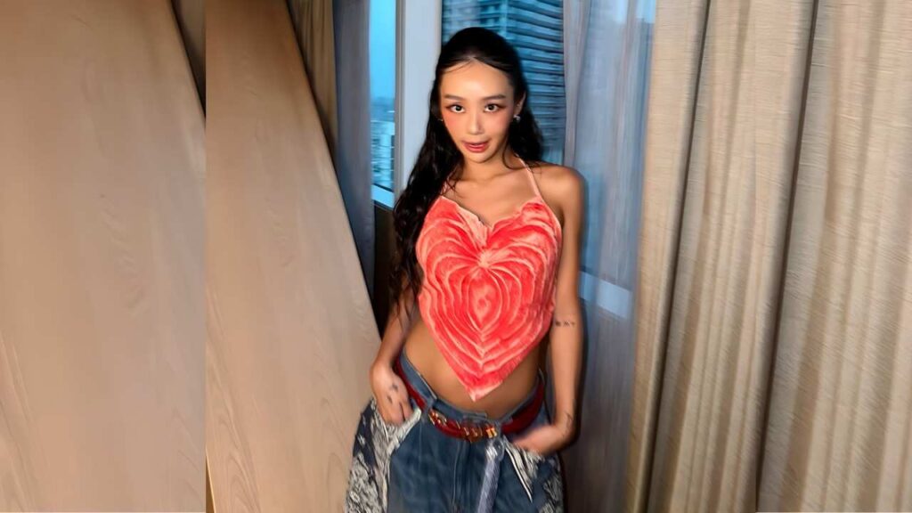 Hyolyn (효린) with long dark hair in a heart-shaped red top and jeans, hands on hips, standing by beige curtains and a window.