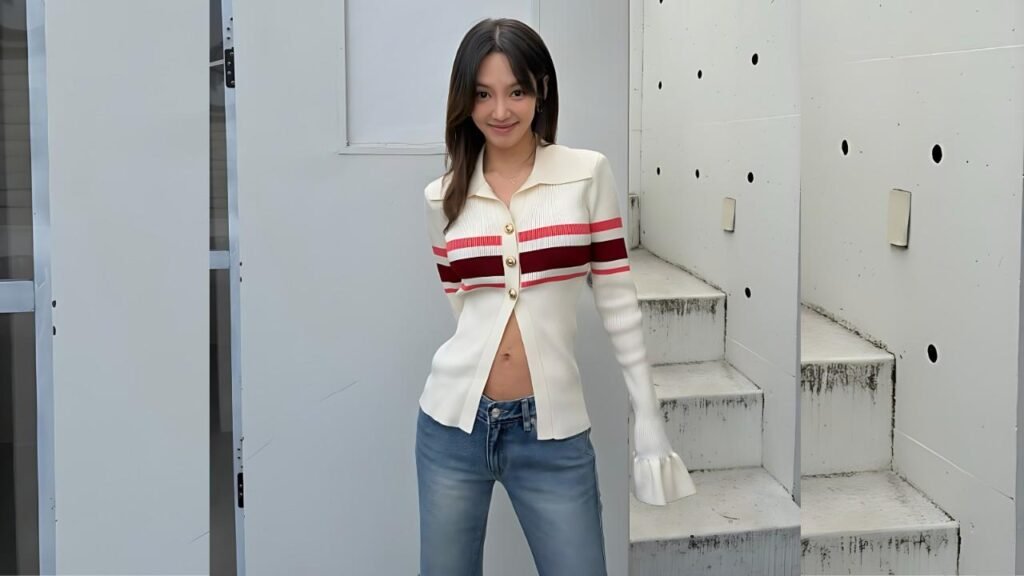 Hyoyeon with dark hair stands outside on stairs, wearing a cream cardigan with red stripes and gold buttons, jeans, smiling.