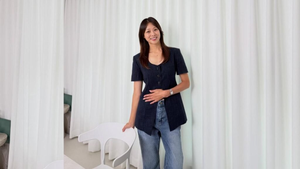 Jang Shin-young in navy blazer and jeans stands before white curtains, one hand on her stomach, the other on a chair.