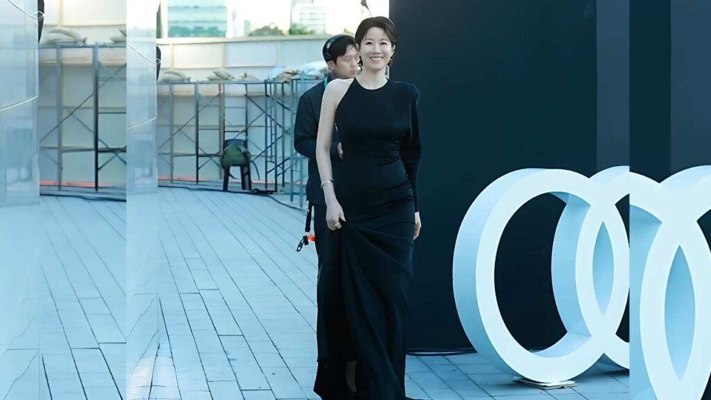 Jeon Hye-jin in a black one-shoulder gown poses on a rooftop beside oversized white interlocking rings, with a man nearby.